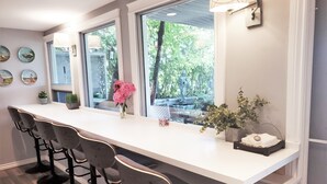 A modern kitchen with a white countertop, four bar stools, a vase with pink flowers, and a view of greenery outside. - Along River Ridge (Calgary)