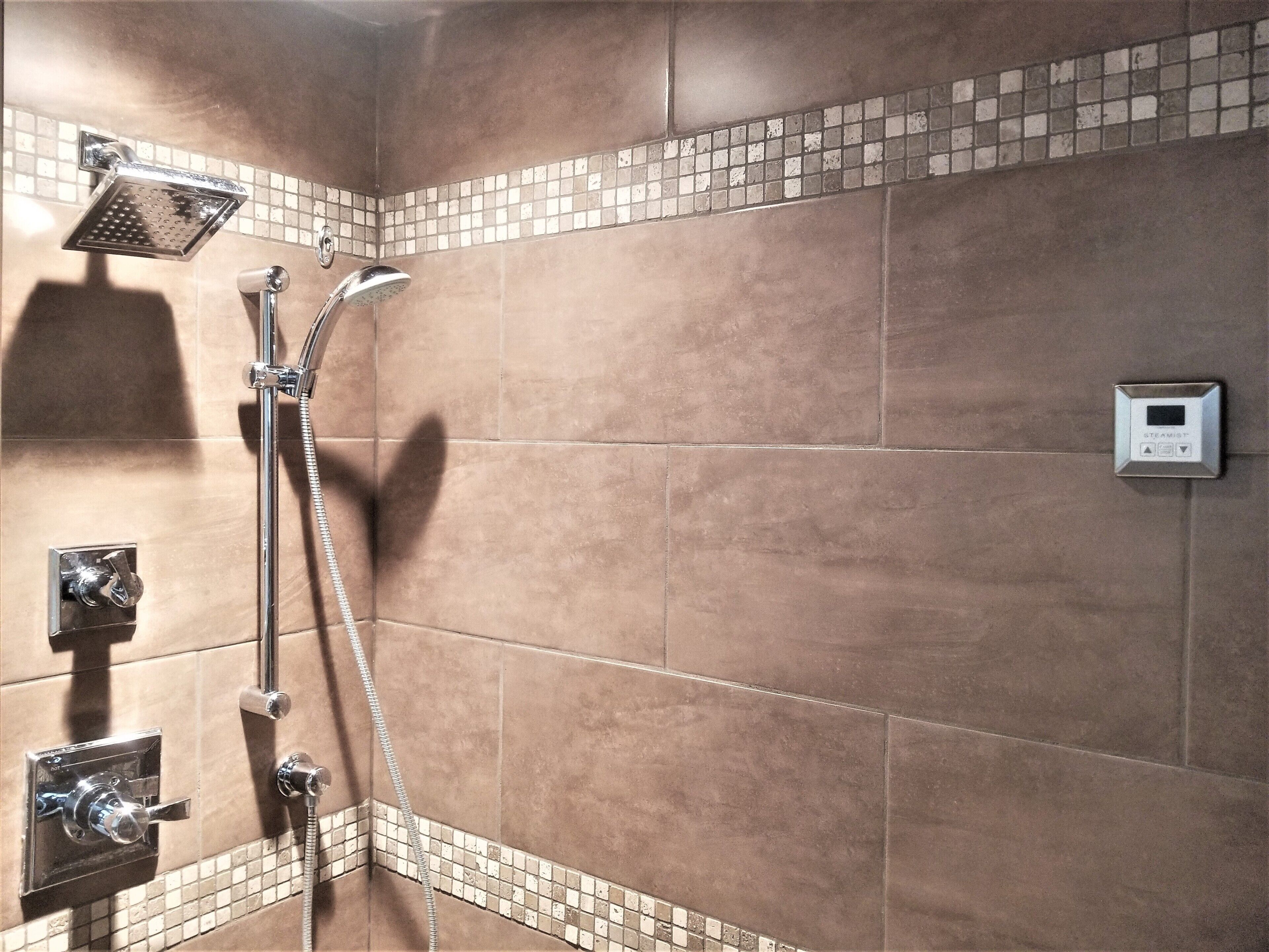 Ducha con cabezal de mano, panel de control en la pared y diseño de azulejos en esquina.