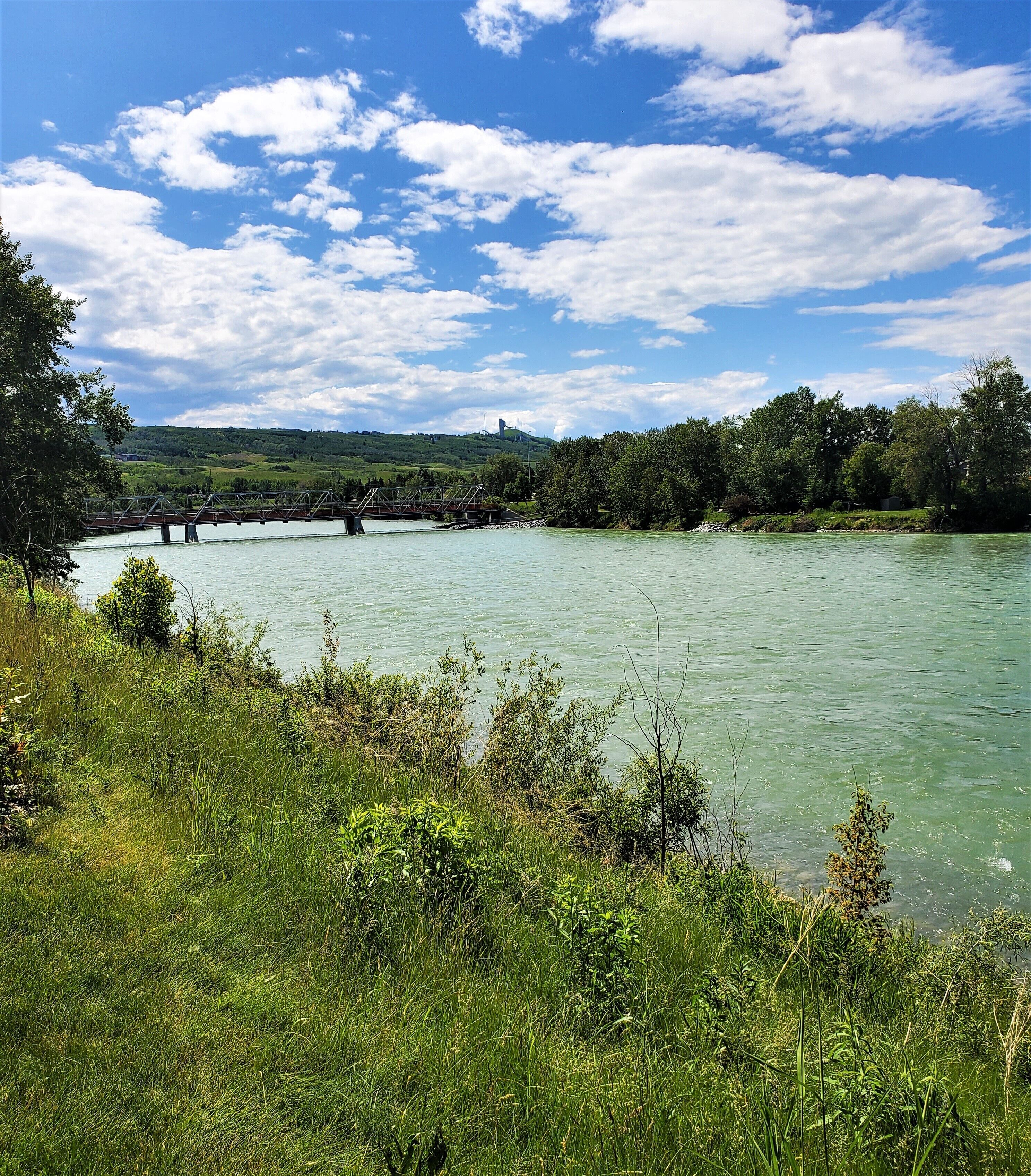 Ein Fluss mit einer Brücke, umgeben von Grünflächen und Hügeln im Hintergrund.
