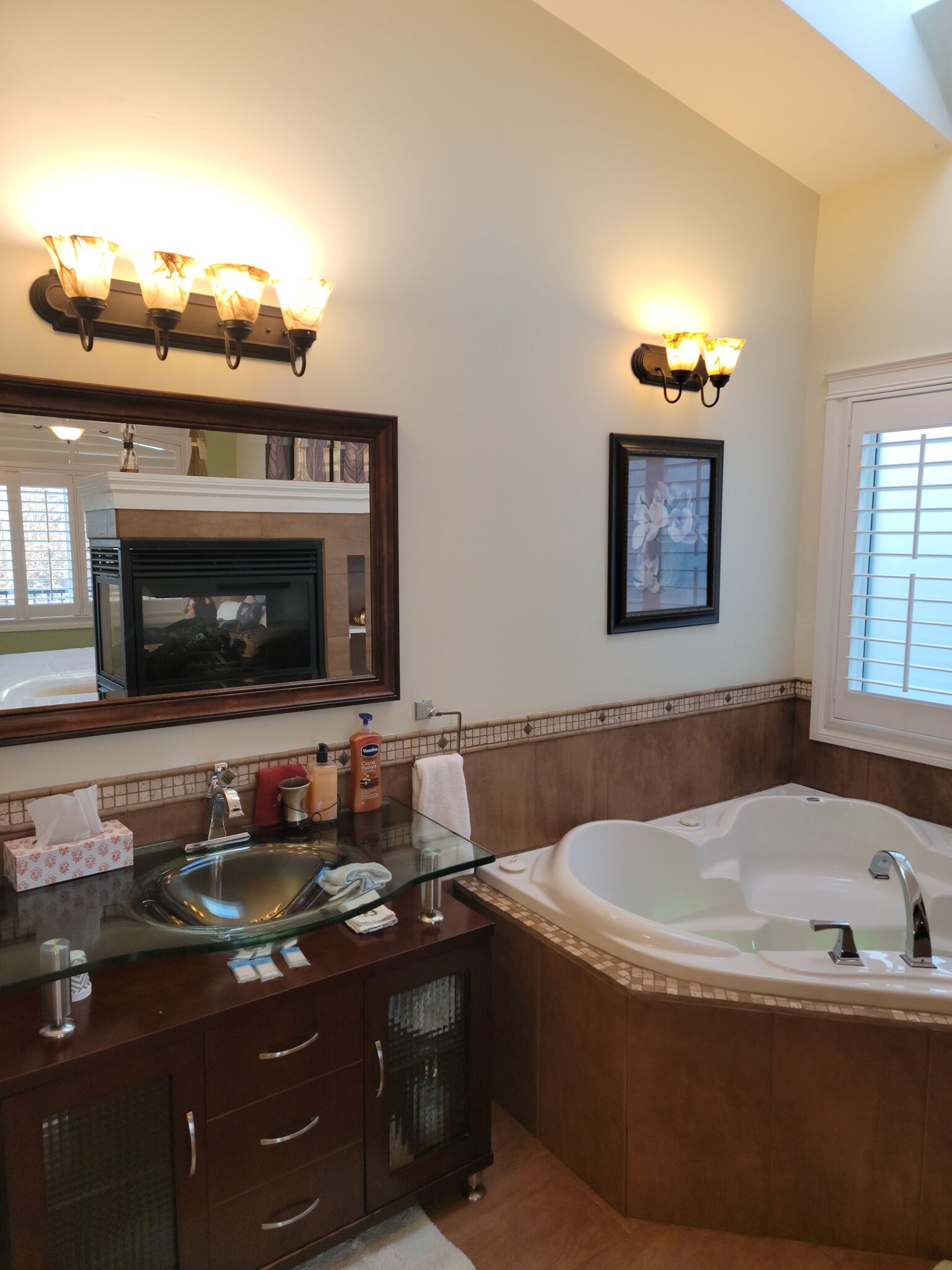 A bathroom with a glass sink, a large bathtub, a mirror with lights, and a framed picture on the wall.