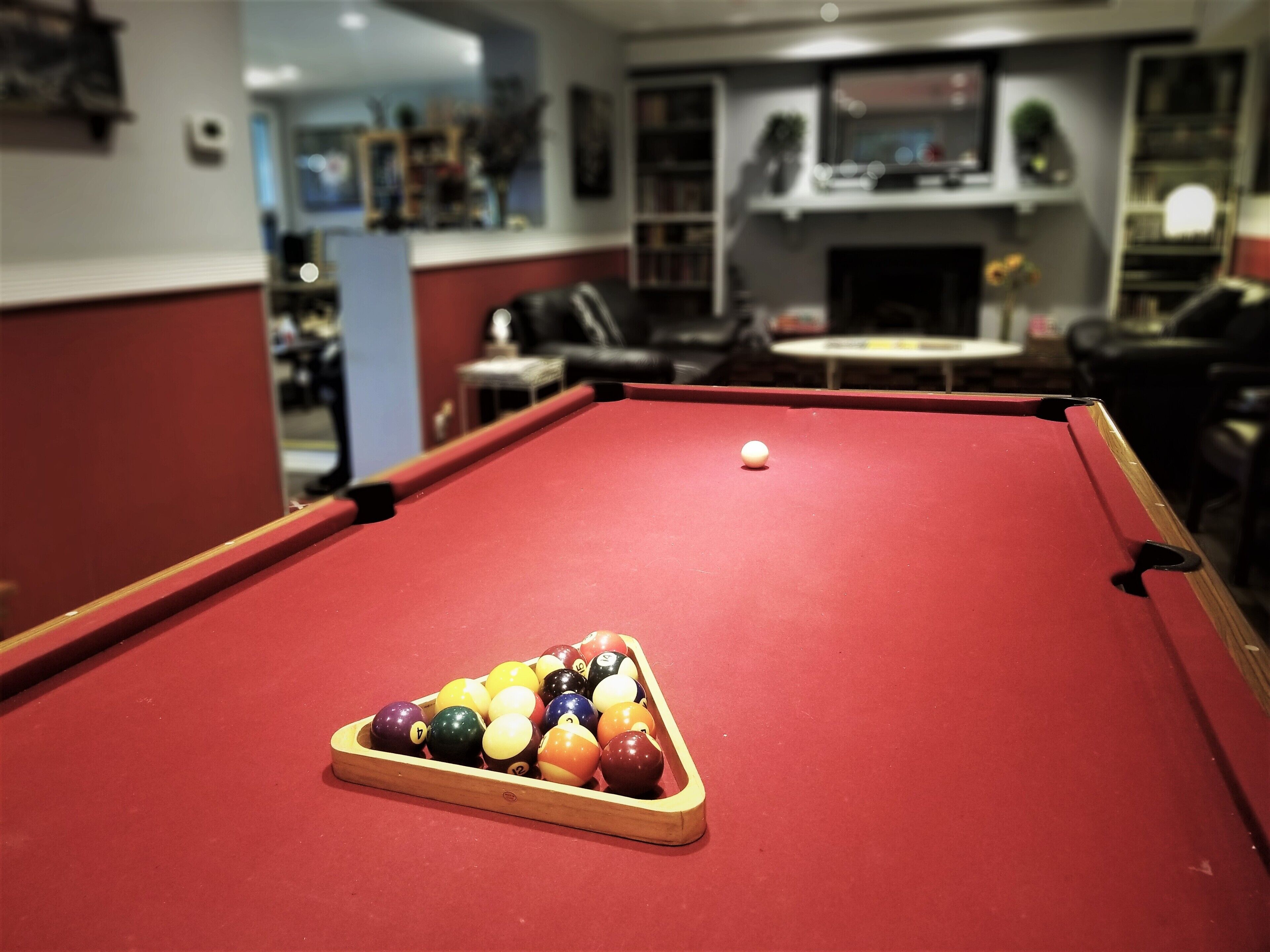 A pool table with a triangular rack of billiard balls in a room with a fireplace and bookshelves.