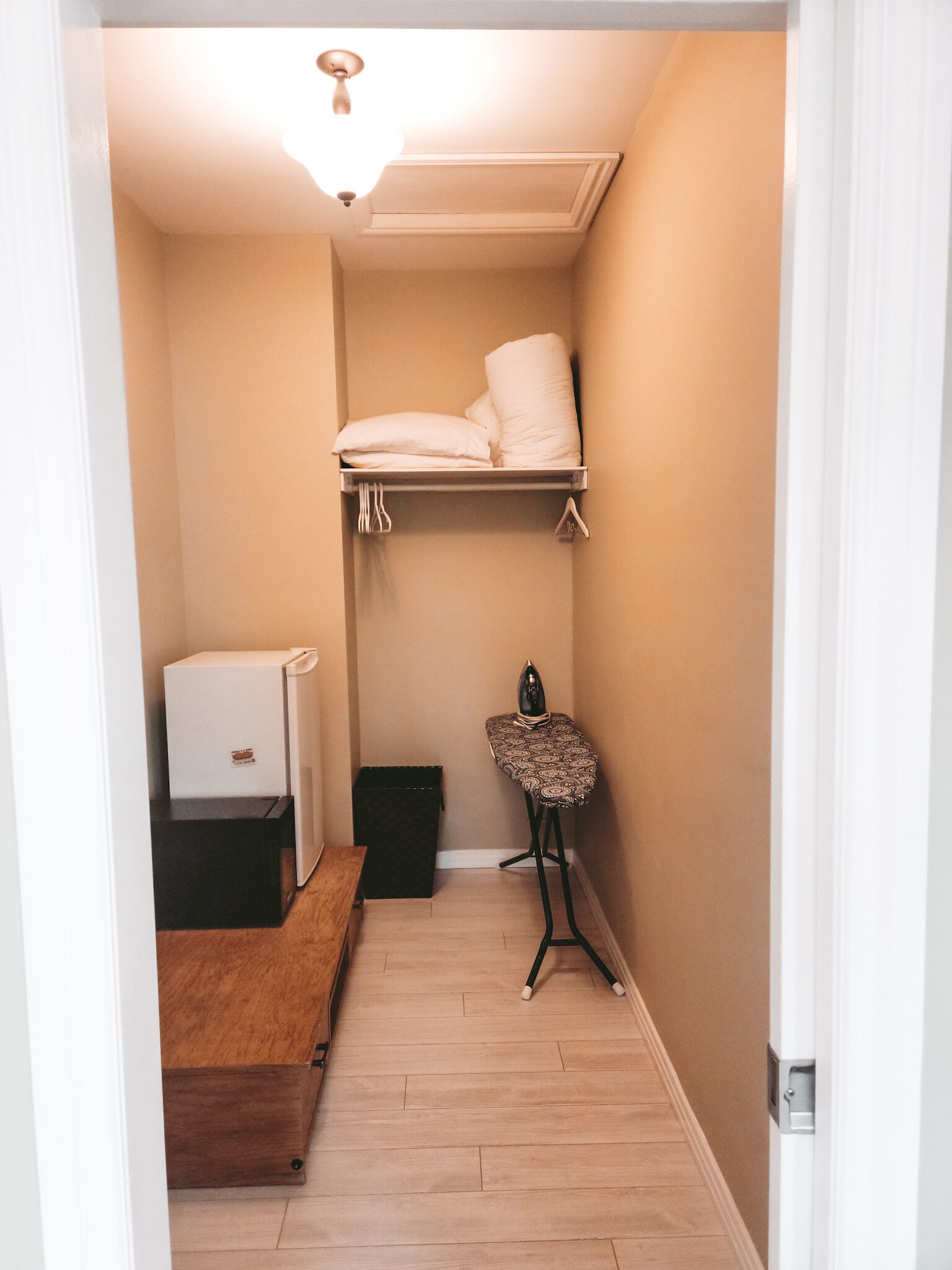 Un pasillo estrecho con una cama individual doble, una plancha pequeña, un refrigerador y un banco de madera.
