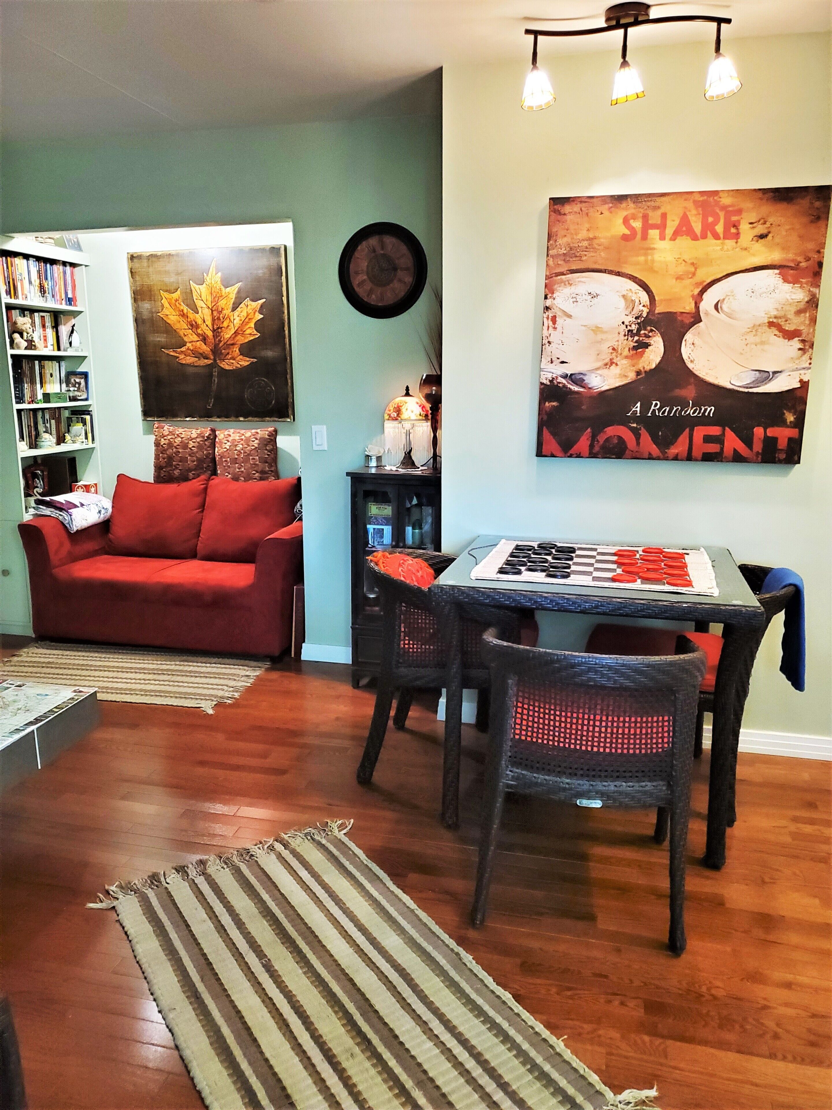 A living room with a red sofa, a dining table with chairs, a bookshelf, and a wall clock.