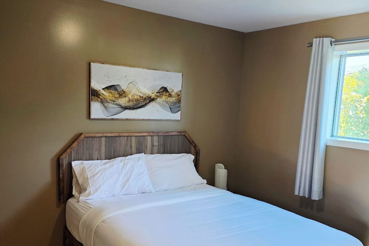 Quarto com estrutura de cama de madeira, roupa de cama branca, luminária no teto, pintura abstrata na parede e janela com cortinas.