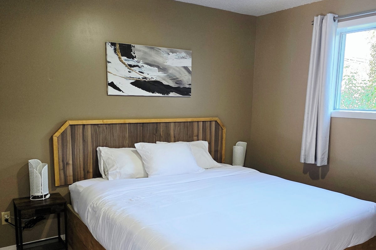 Quarto com cama de madeira, mesa de cabeceira, luminária na parede e um quadro.