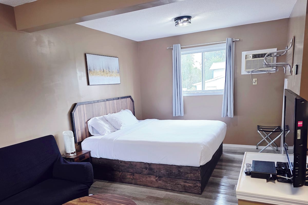 Quarto de hotel com uma cama, um sofá escuro, uma pequena mesa de madeira, uma TV e uma janela com cortinas.
