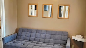 A living room with a grey sofa, a black coffee table, and a wooden side table. There are three mirrors on the wall and a ceiling light fixture.