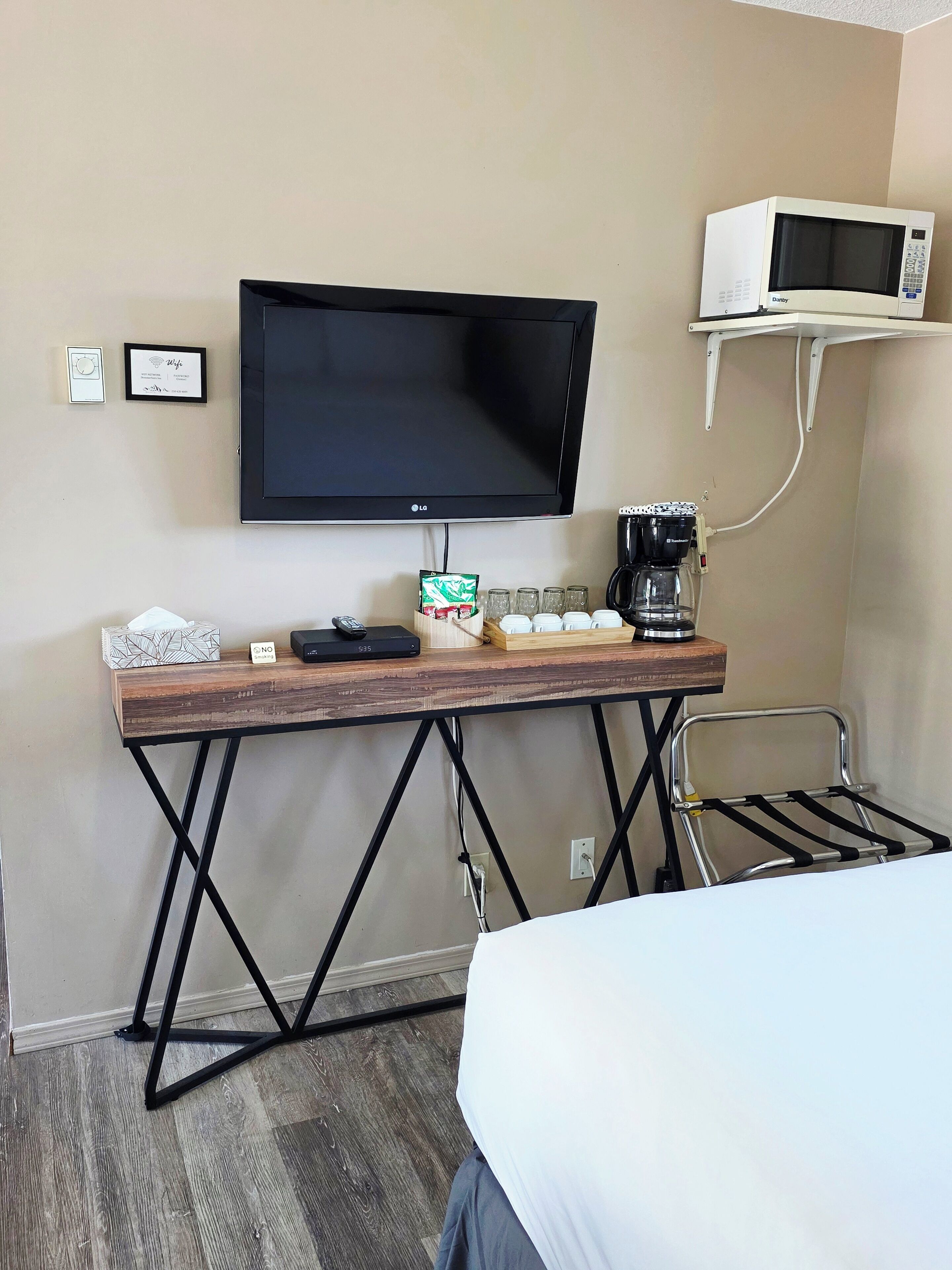 Habitación de hotel con televisor de pantalla plana en la pared, cafetera, una mesita con control remoto y una silla con perchero.