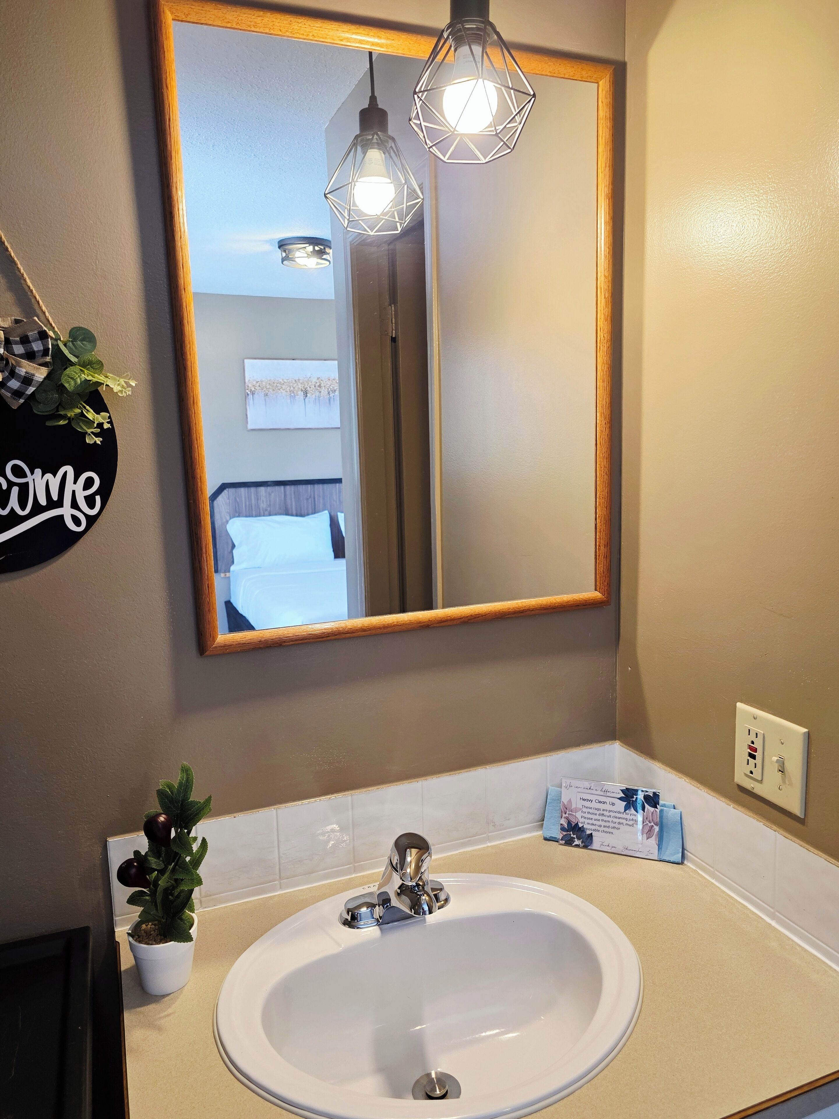 A bathroom with a large mirror, a white sink, a potted plant, and a welcome sign.