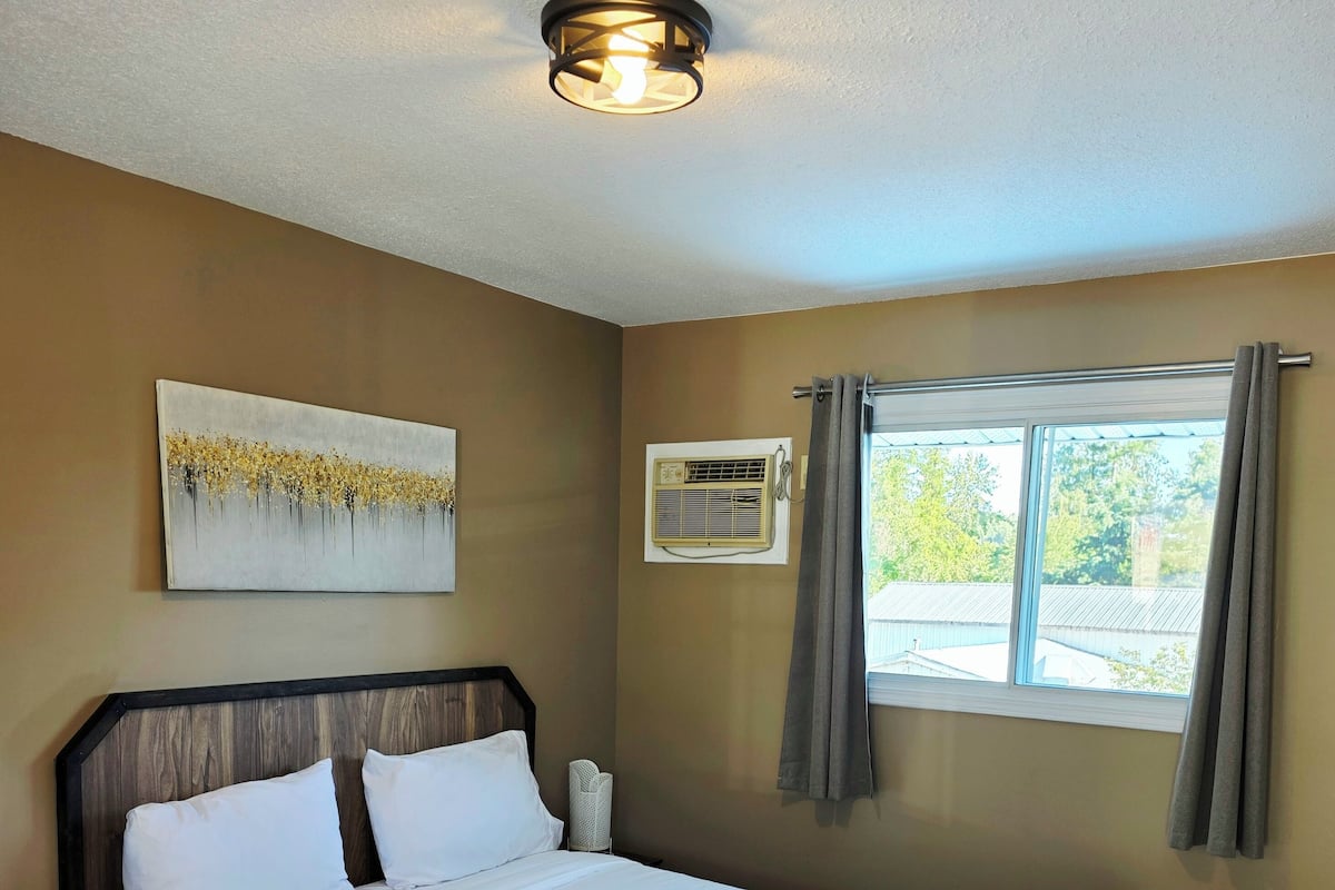 Cama bem arrumada em quarto com piso de madeira, luminária no teto, janela com cortinas e ar-condicionado fixado na parede.