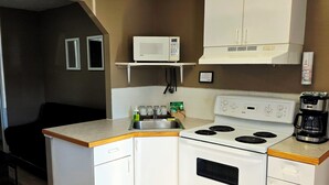 A compact kitchen with white appliances, a microwave, a stove, and a coffee maker. There is a wooden table and a small countertop.