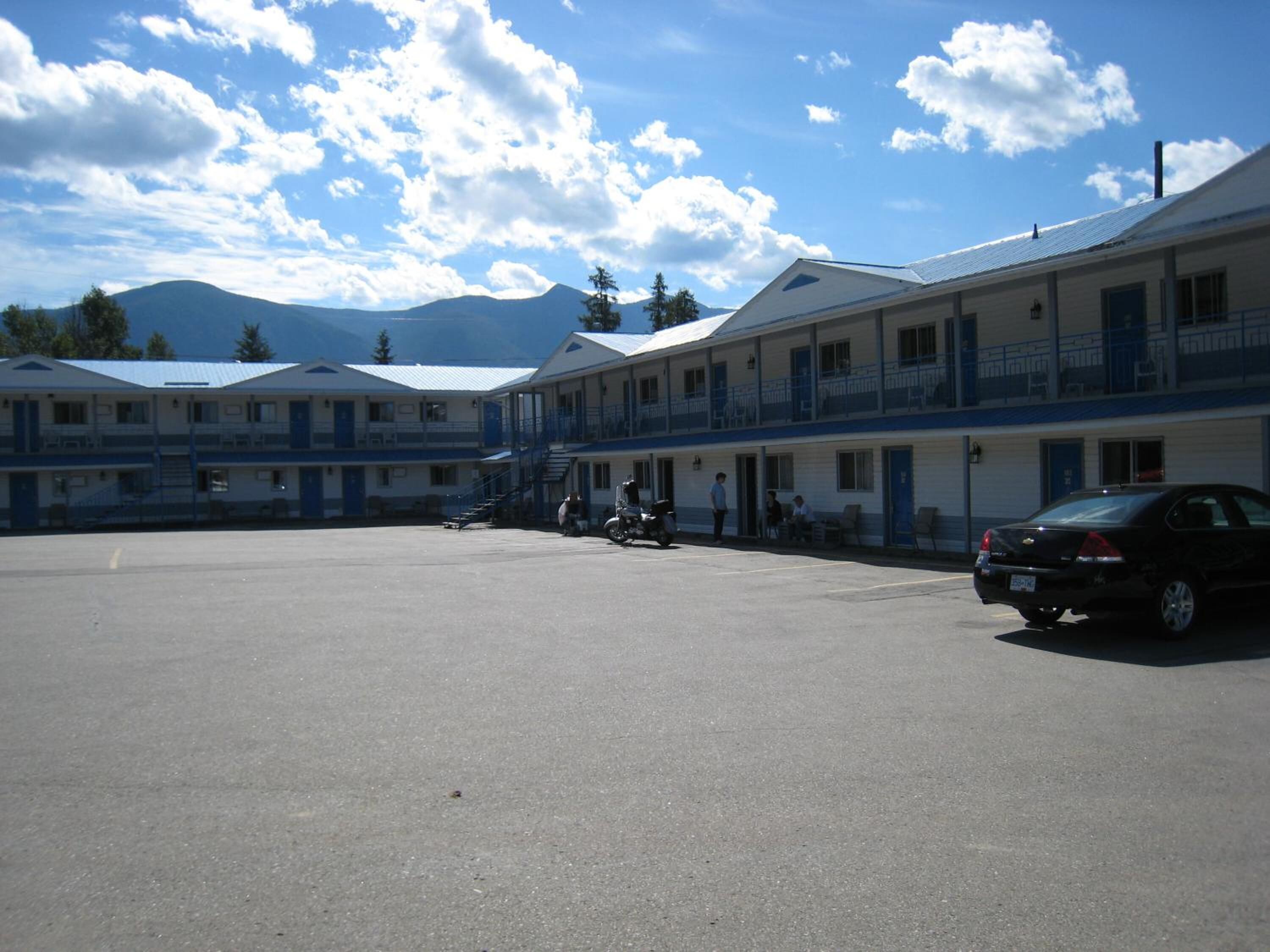Un motel con varias habitaciones, un estacionamiento y montañas al fondo.