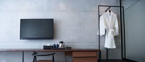 A modern hotel room with a flat-screen TV, a wooden desk, a chair, a robe hanging on a rack, and a coffee maker.
