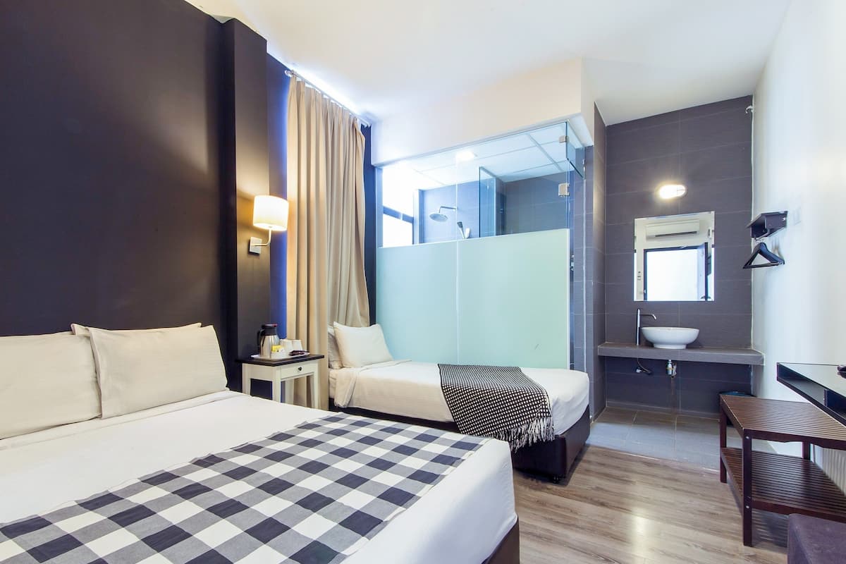 Habitación de hotel con una cama grande, una mesita de noche, una silla, un escritorio pequeño y un baño visible a través de un panel de vidrio.