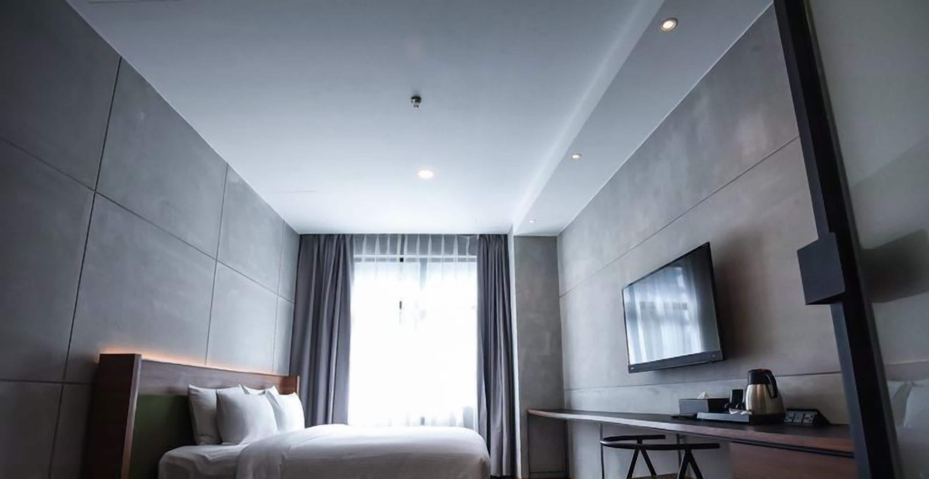 A modern hotel room with a large bed, a flat-screen TV, a small table with a coffee maker, and a large window with curtains.