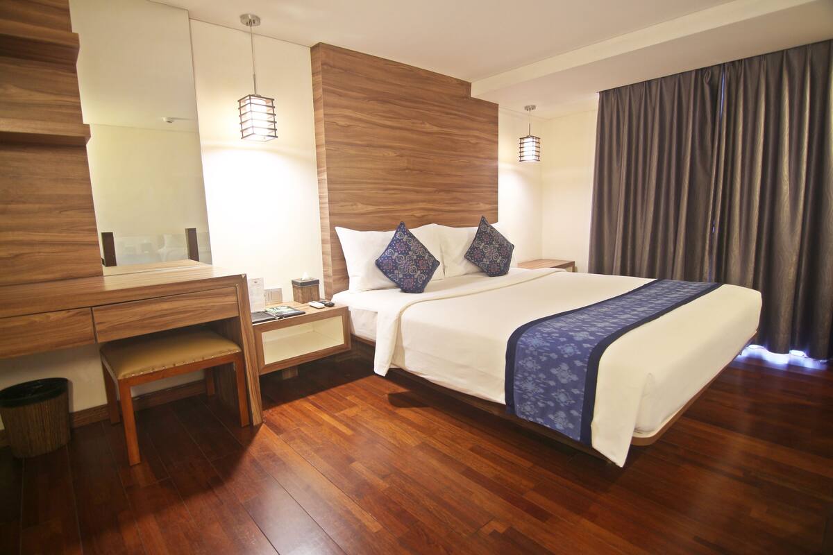 A hotel room with a wooden floor, a bed with white bedding and blue pillows, a desk with a chair, a nightstand, and a large mirror.