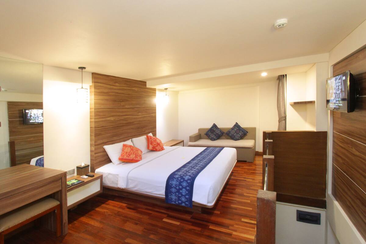 A hotel room with a wooden floor, a bed with white bedding and orange pillows, a wooden desk, a small table, a sofa with blue cushions, and a TV mounted on the wall.
