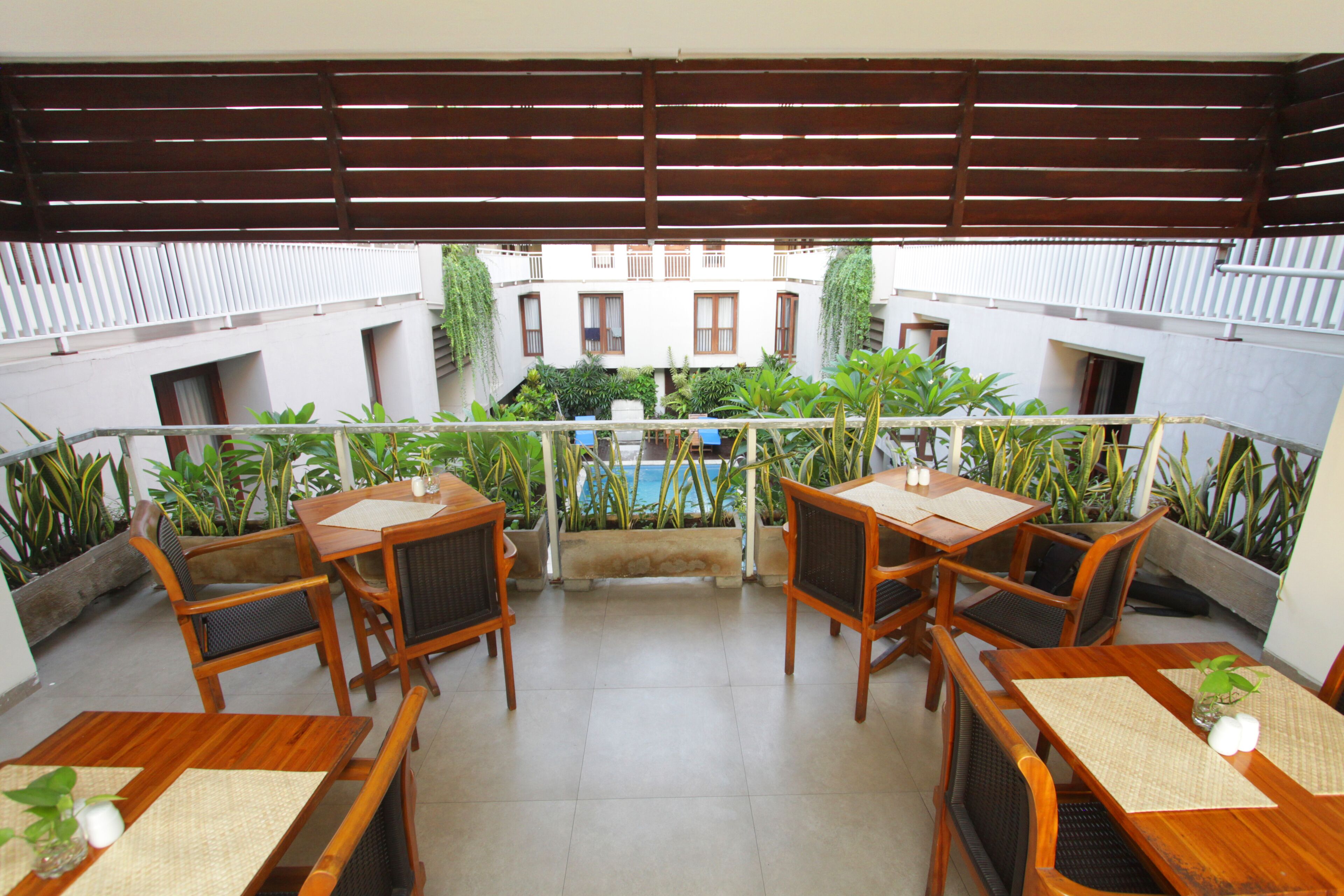 A balcony with wooden furniture and potted plants, overlooking a courtyard with a pool and buildings.