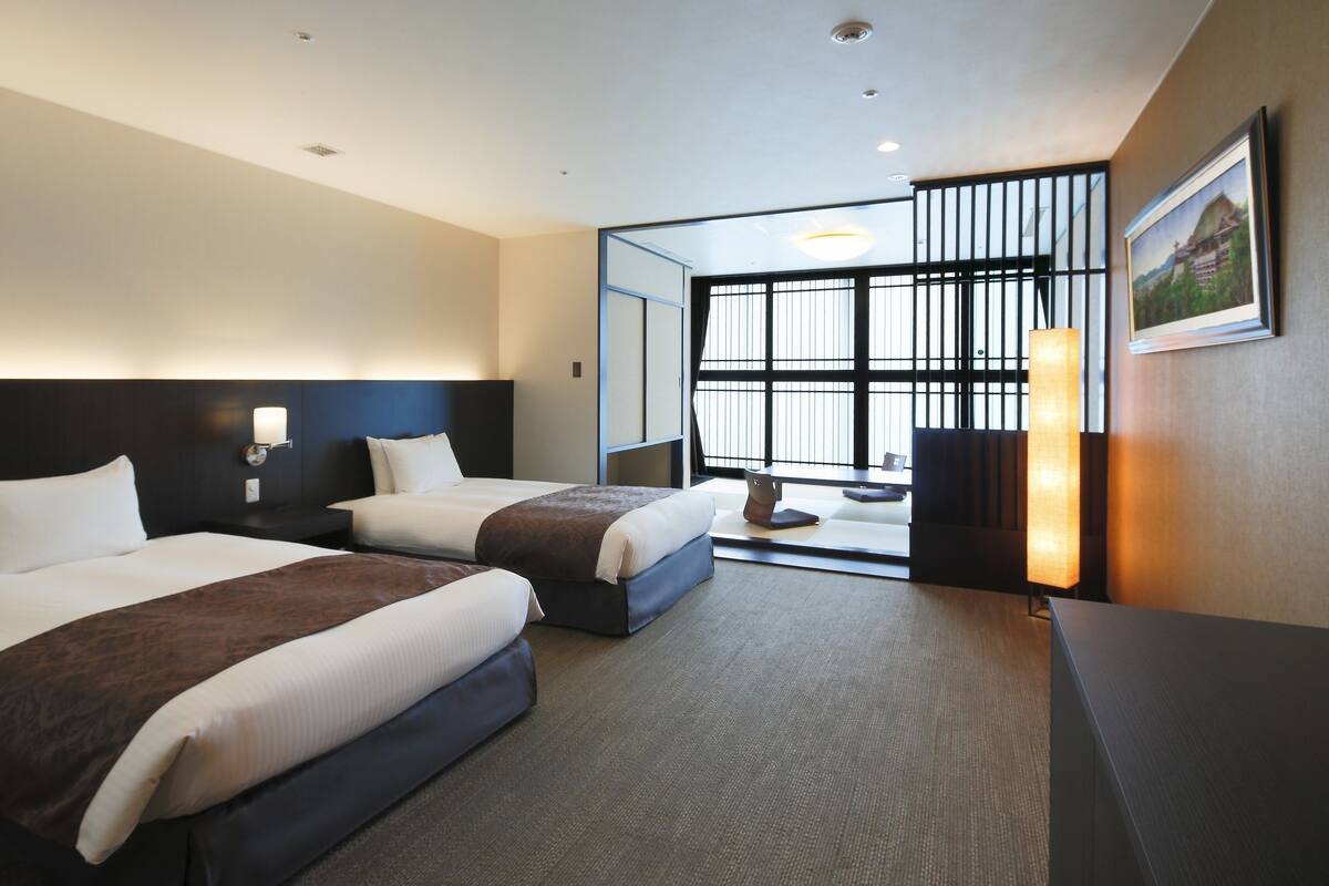 A hotel room with two beds, a desk, a chair, a lamp, and a large window with blinds.
