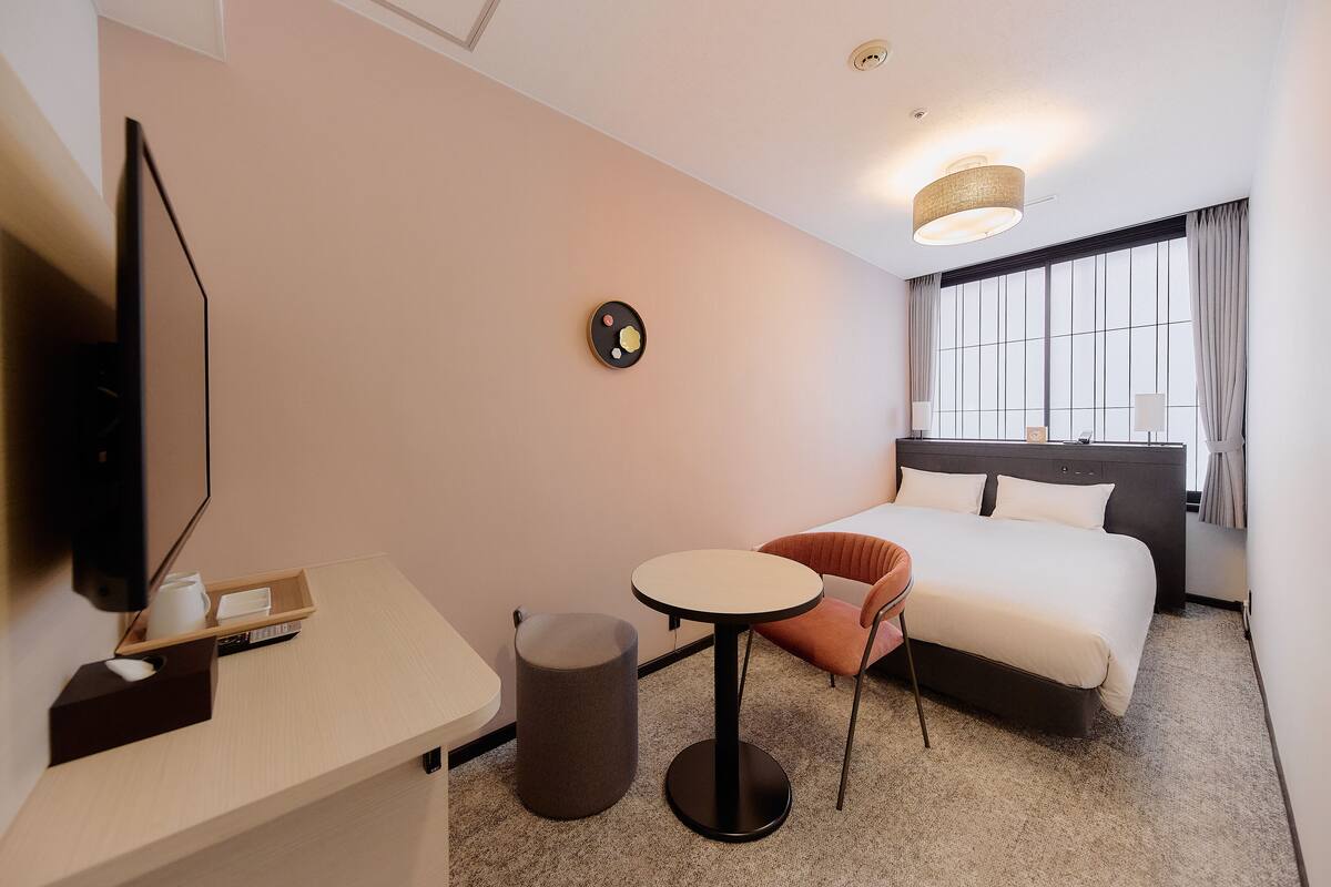 A hotel room with a bed, a desk with a computer, a chair, a round table, and a trash bin.