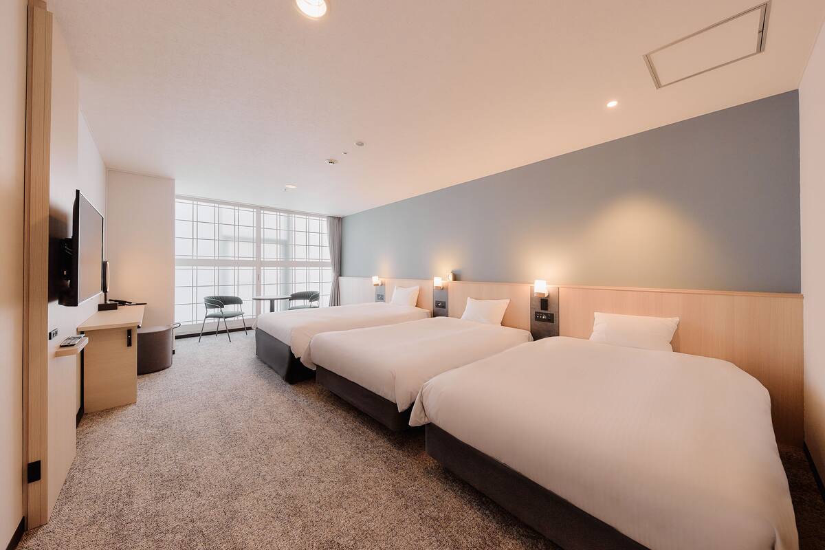 A hotel room with three beds, a desk, a TV, and a large window.