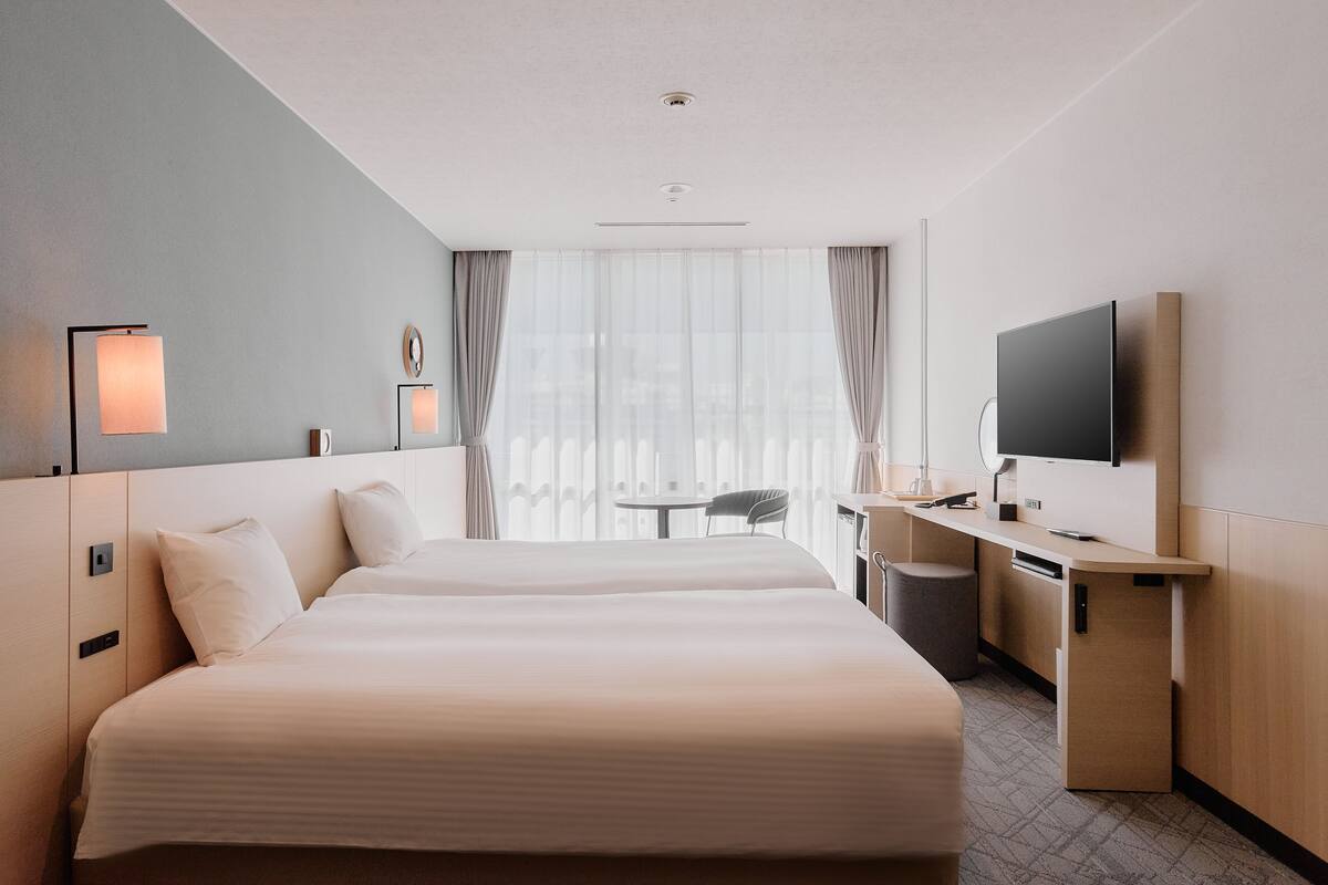 A hotel room with two beds, a desk, a chair, a TV, and a window with curtains.