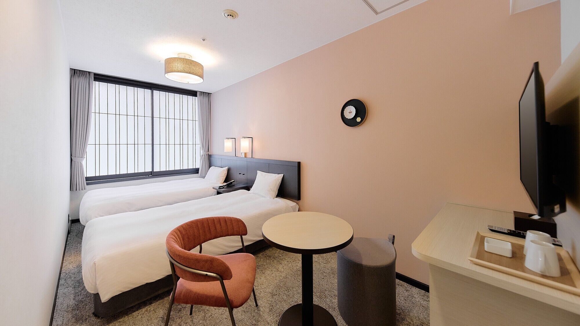 Hotellihuone, jossa on kaksi sänkyä, pieni pyöreä pöytä, tuoli, televisio ja seinällä oleva kello.