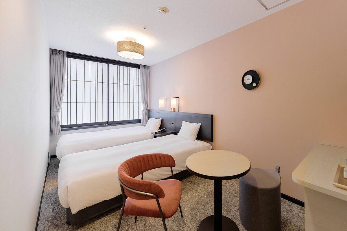 A hotel room with two beds, a small round table, a chair, a TV, and a clock on the wall.