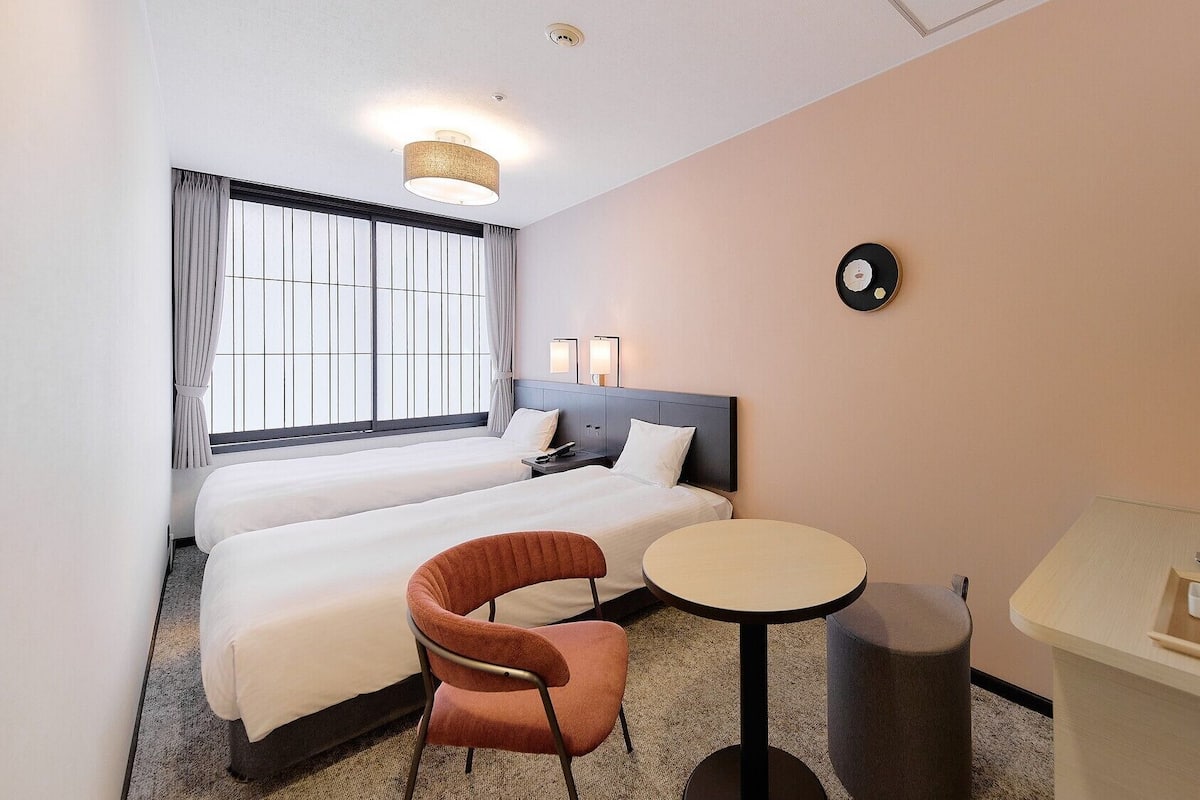 Hotellihuone, jossa on kaksi sänkyä, pieni pyöreä pöytä, tuoli, televisio ja seinällä oleva kello.