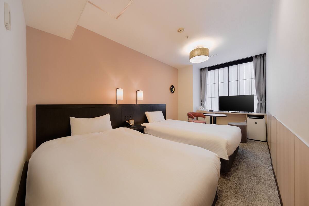 A hotel room with two beds, a desk, a television, and a window with curtains.