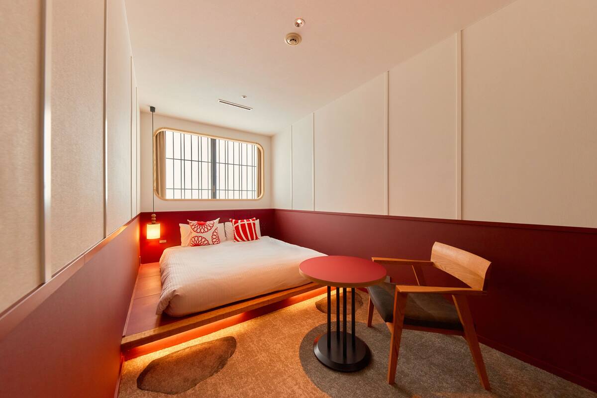 A compact hotel room with a bed, a small round table, a chair, and a window with barred glass.