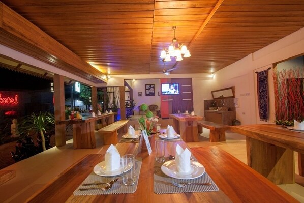 A restaurant with wooden tables set for dining, a bar area, and a television screen in the background.