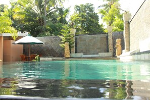 A swimming pool with a stone wall, a statue, and a chair under an umbrella. - Green Asri (Senggigi)