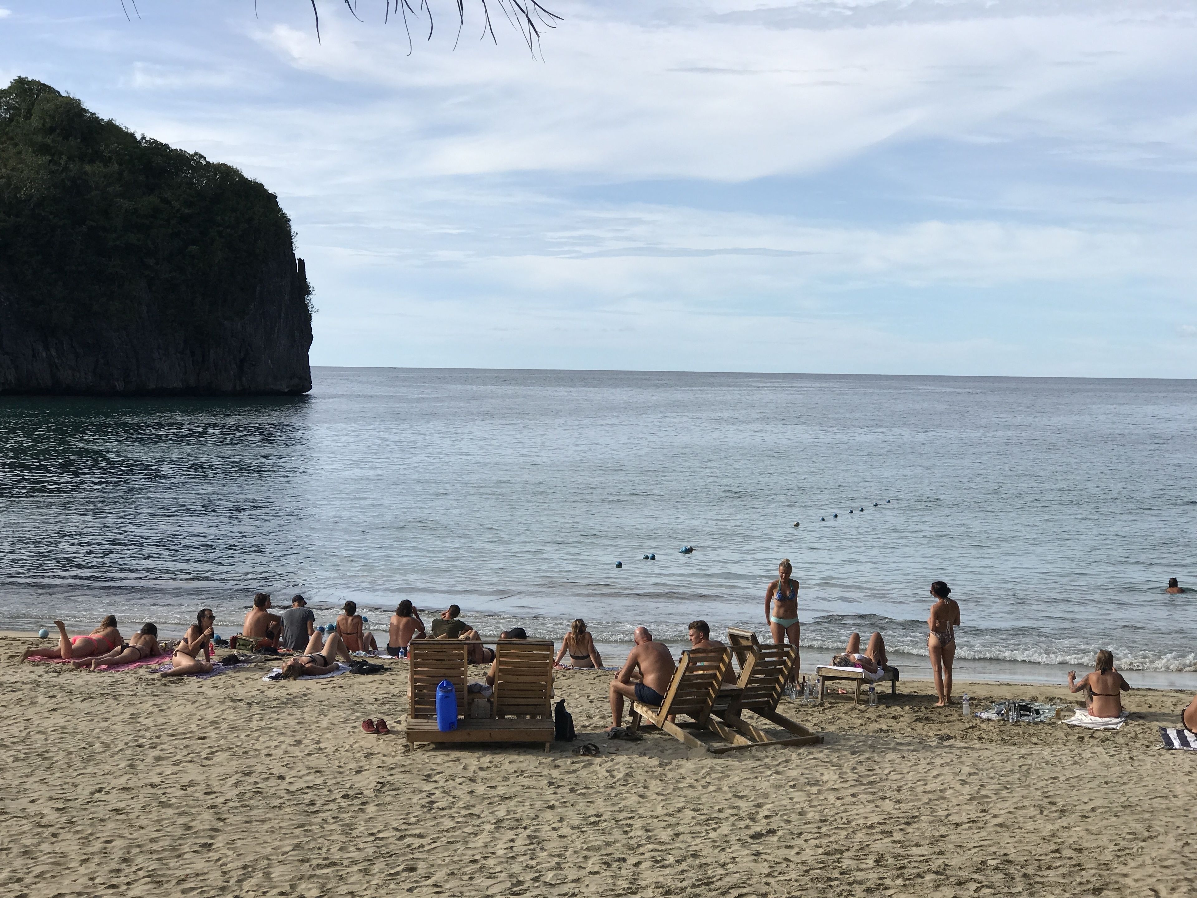 A beach with people sunbathing and swimming, wooden chairs, and a cliff in the background.