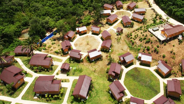 Vista aérea de un resort con varias cabañas dispuestas en un círculo alrededor de un césped central, con una playa y colinas boscosas al fondo.