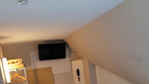 Quarto compacto com escrivaninha, duas cadeiras, uma mesinha, TV fixada na parede e um guarda-roupa com cabideiro.