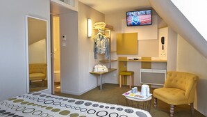 Quarto de hotel com uma cama, uma poltrona amarela, uma mesinha com bandeja, uma televisão na parede e uma porta de banheiro.