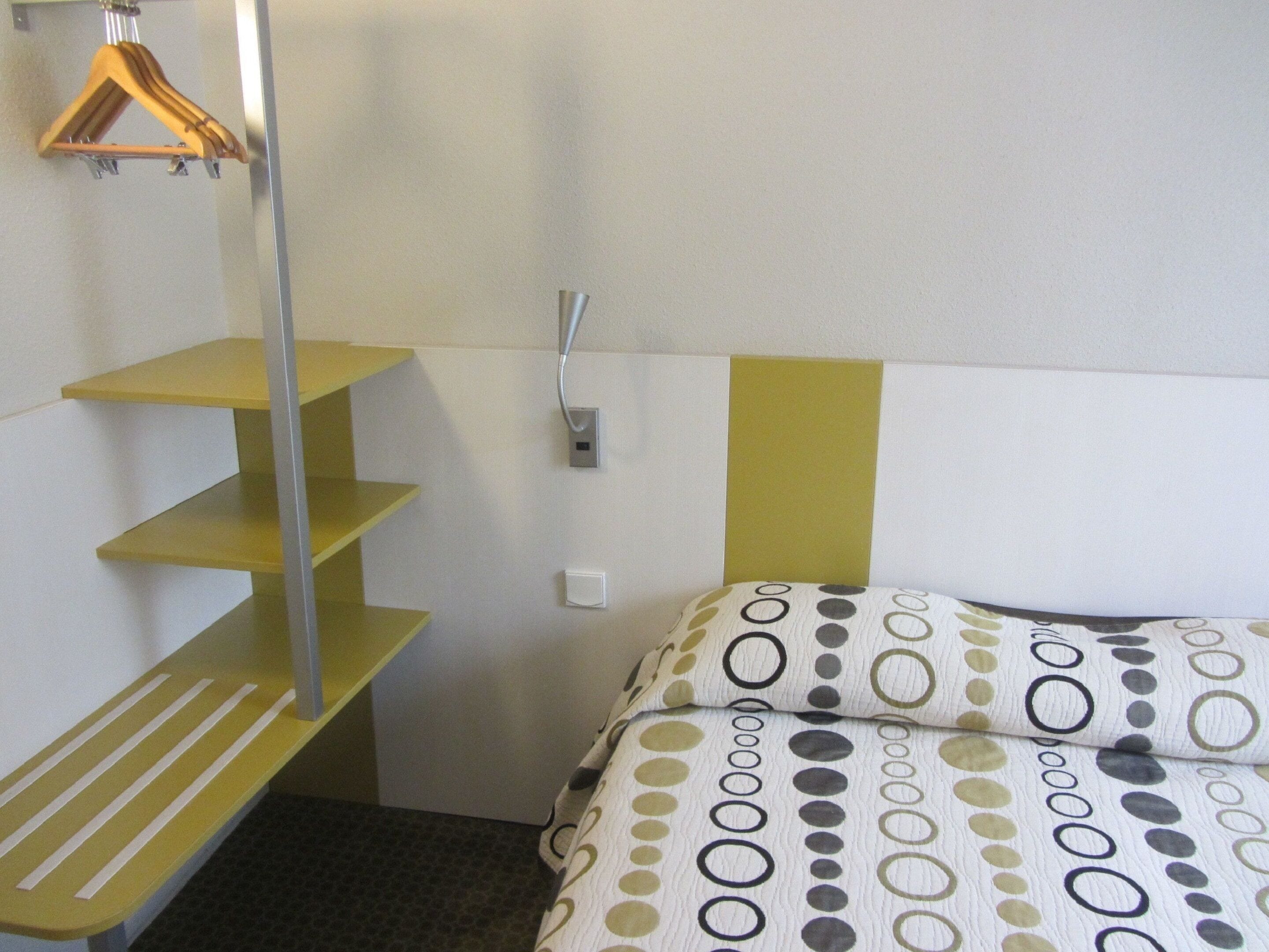 A small, compact room with a bed, a bench, a wall-mounted shelf, and a wall socket.