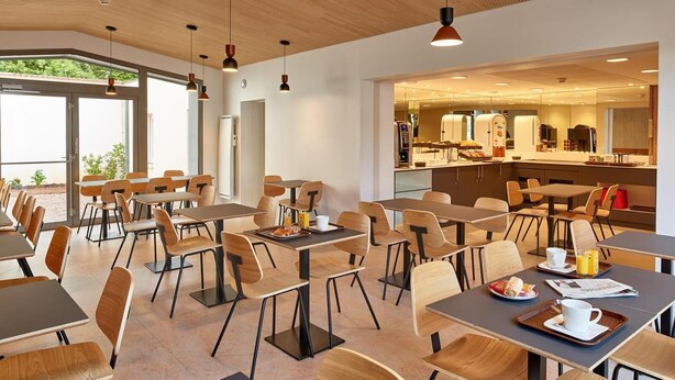 Uma área de refeições moderna com mesas e cadeiras de madeira, uma cozinha ao fundo e um teto com padrão em madeira.