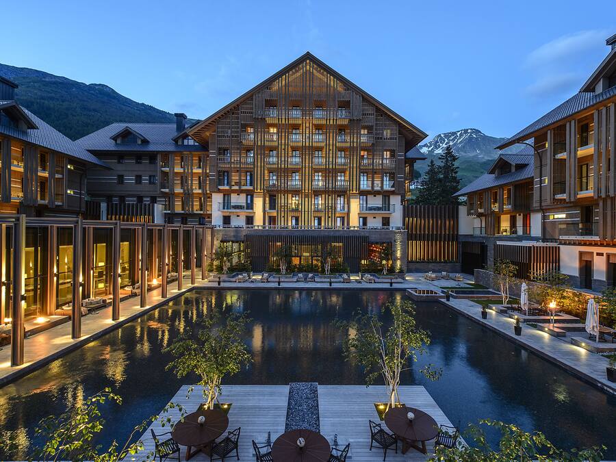Un hotel con piscina central, varios balcones y montañas al fondo.