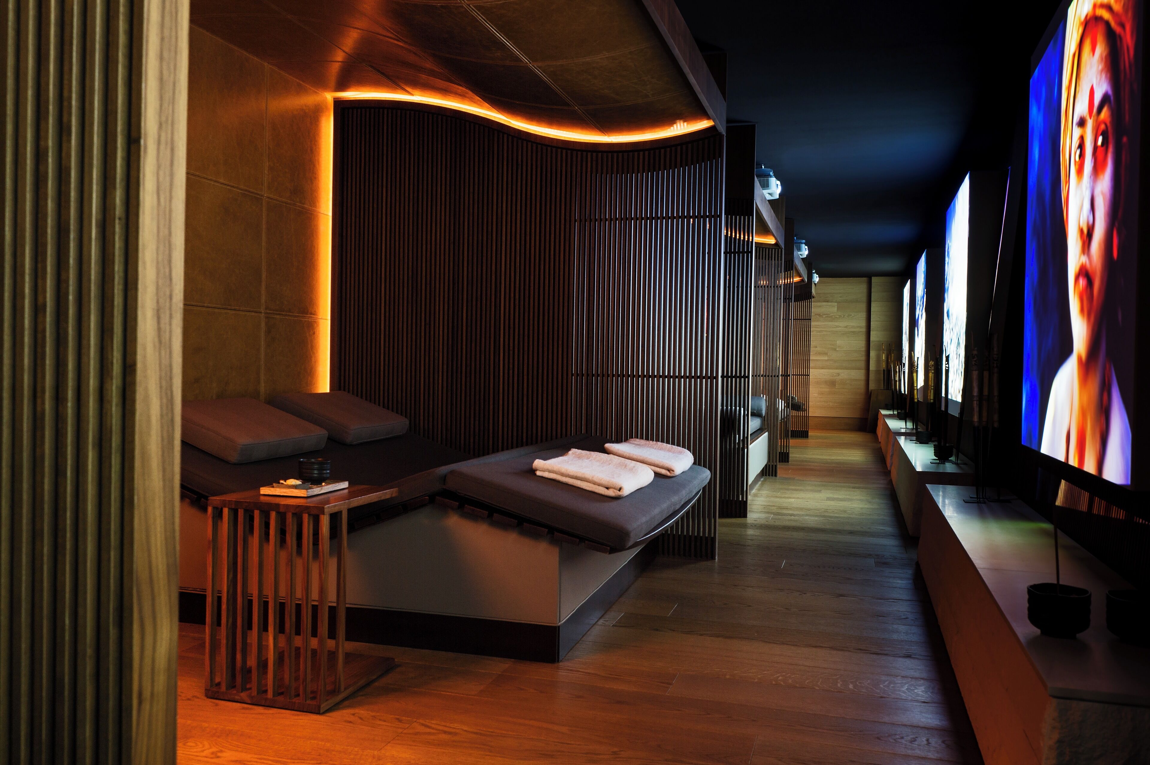 A modern spa room with a massage table, wooden paneling, and a large screen displaying a person.
