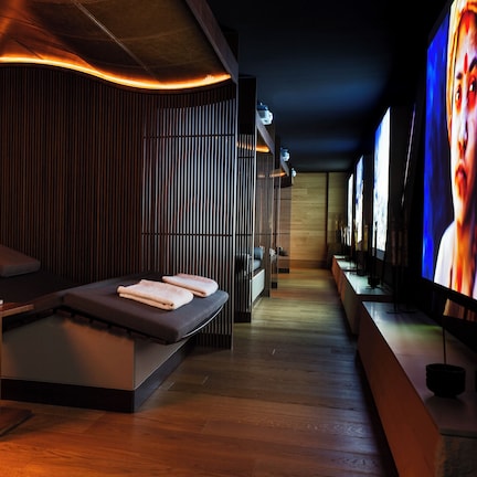 A modern spa room with a massage table, wooden paneling, and a large screen displaying a person.