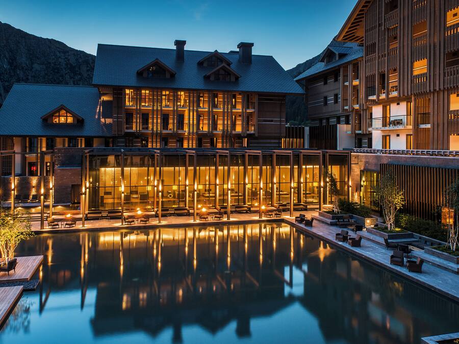 Un hotel moderno con una gran piscina, mobiliario exterior y el paisaje de montañas al fondo.