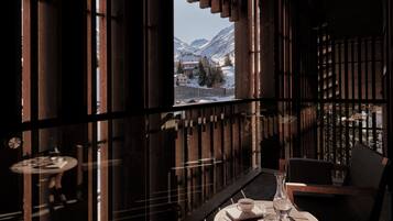 Una habitación acogedora con detalles de madera, una mesa redonda con un florero, una taza y un libro, y vistas a montañas nevadas a través de una ventana.