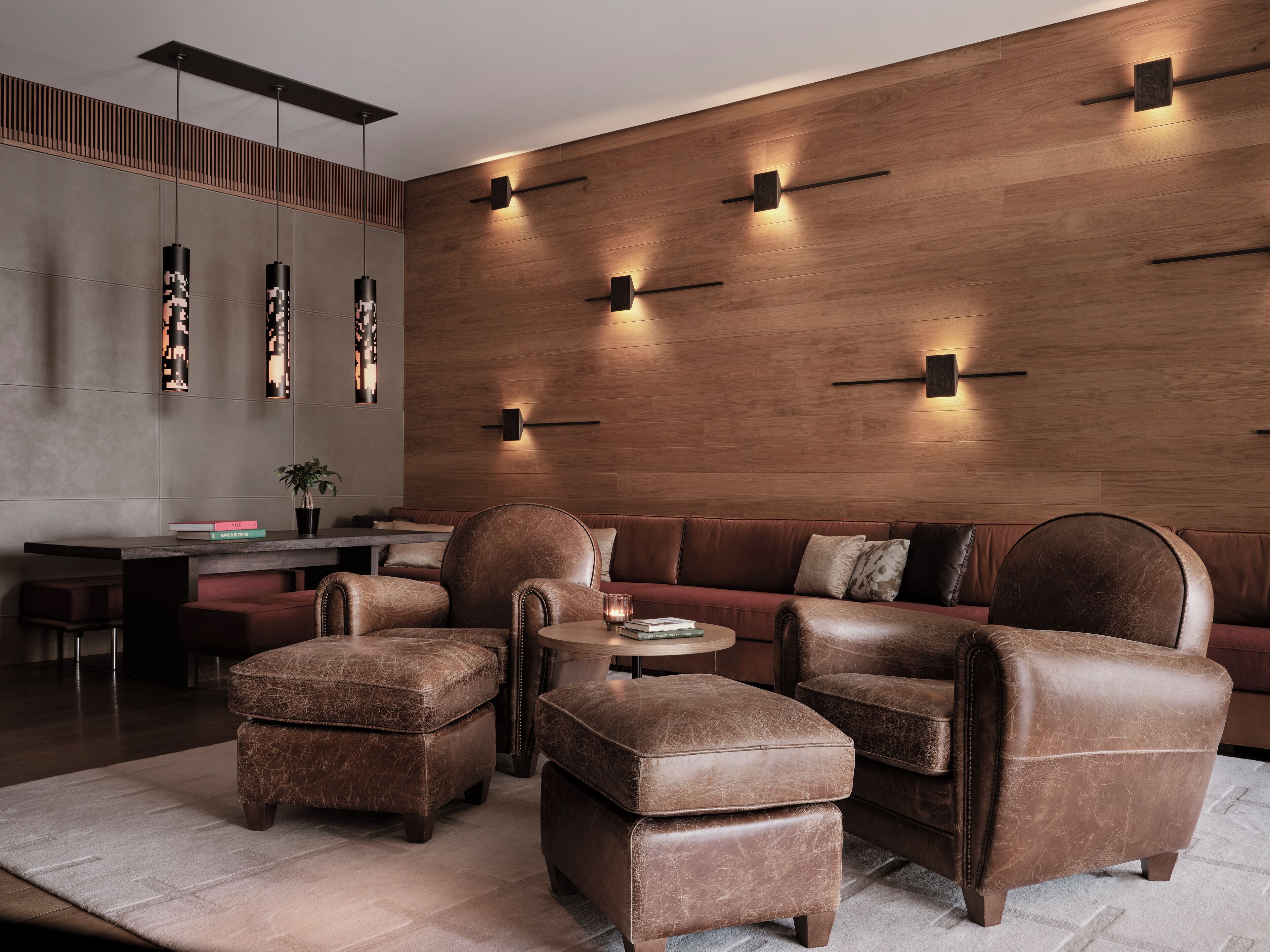 A modern living room with a leather sofa, leather armchairs, and ottomans. There is a small round table with a book and a candle. The room features a wooden accent wall with built-in lighting and a grey accent wall with a vertical light fixture.