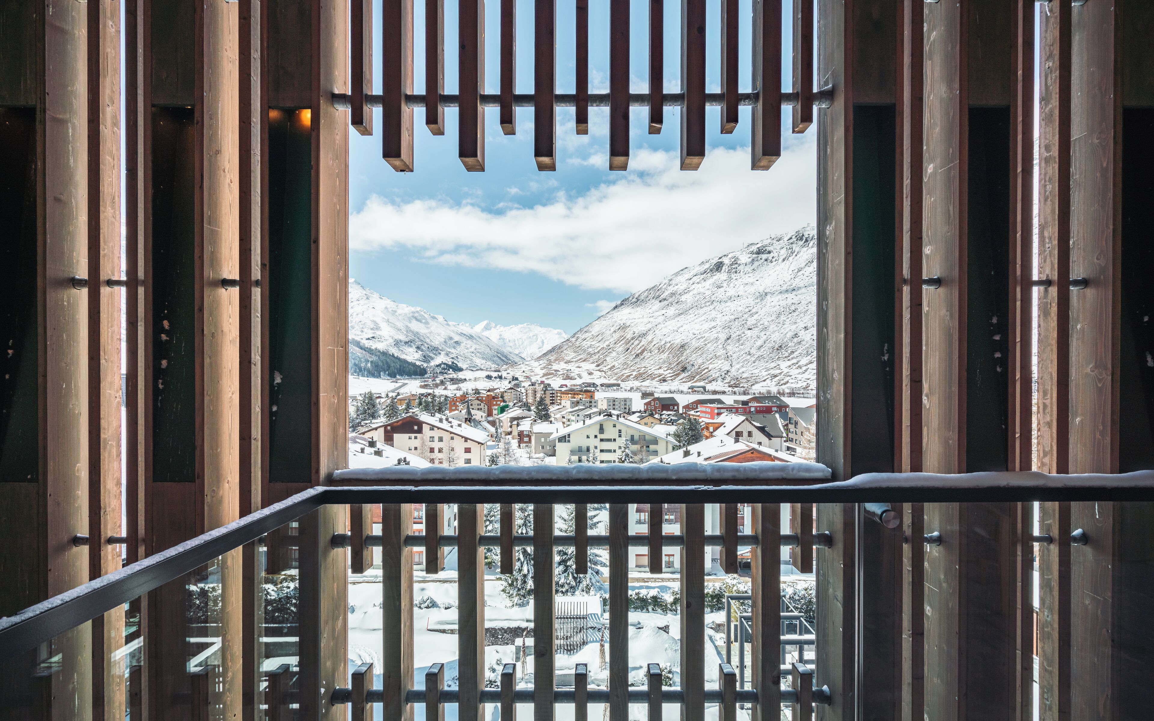 A view from a wooden balcony with metal railings, overlooking a snow-covered town with buildings and mountains in the background.