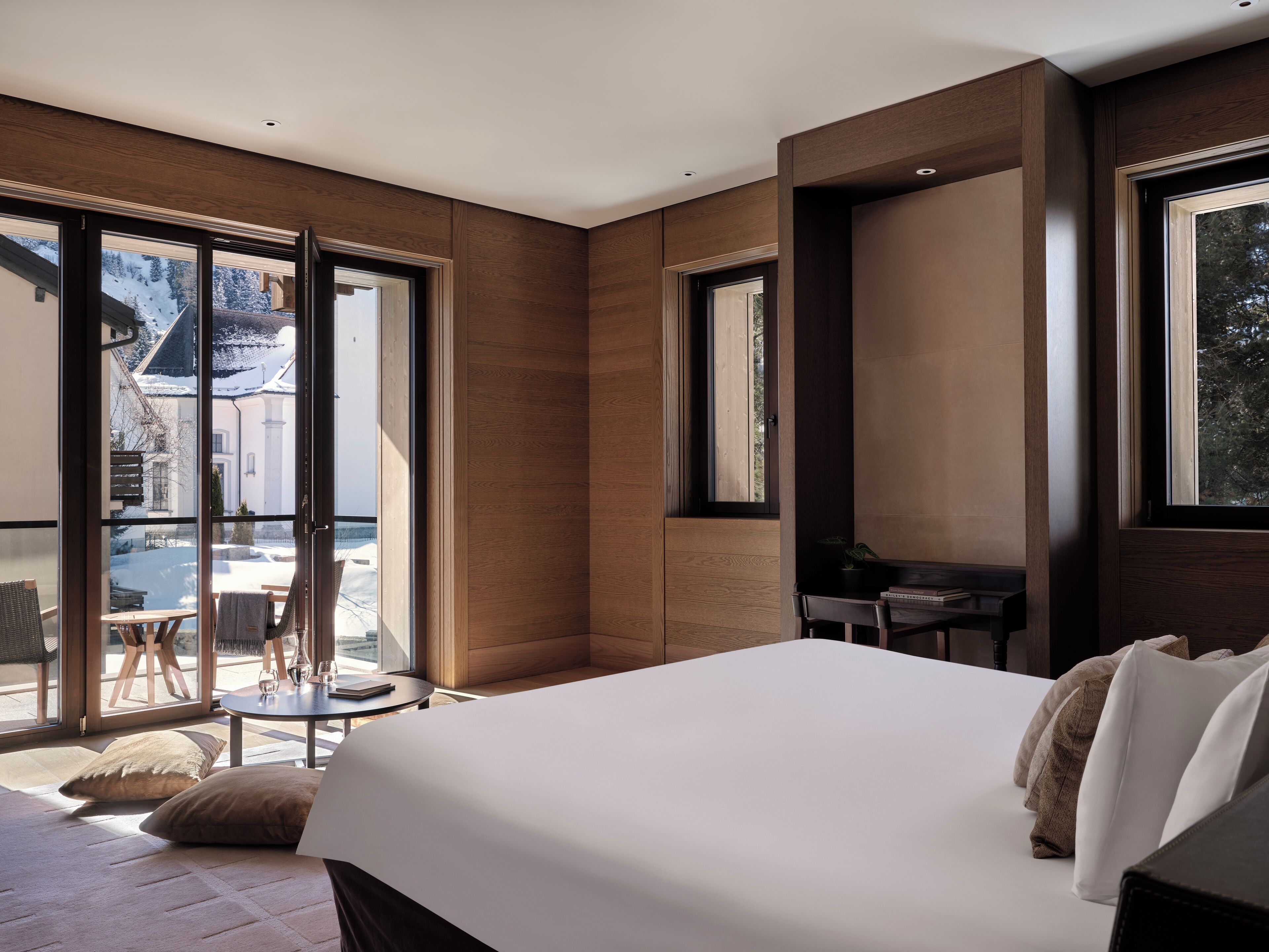 Une chambre d’hôtel moderne, dotée d’un grand lit, d’un petit bureau et d’un balcon offrant une vue sur un paysage enneigé.