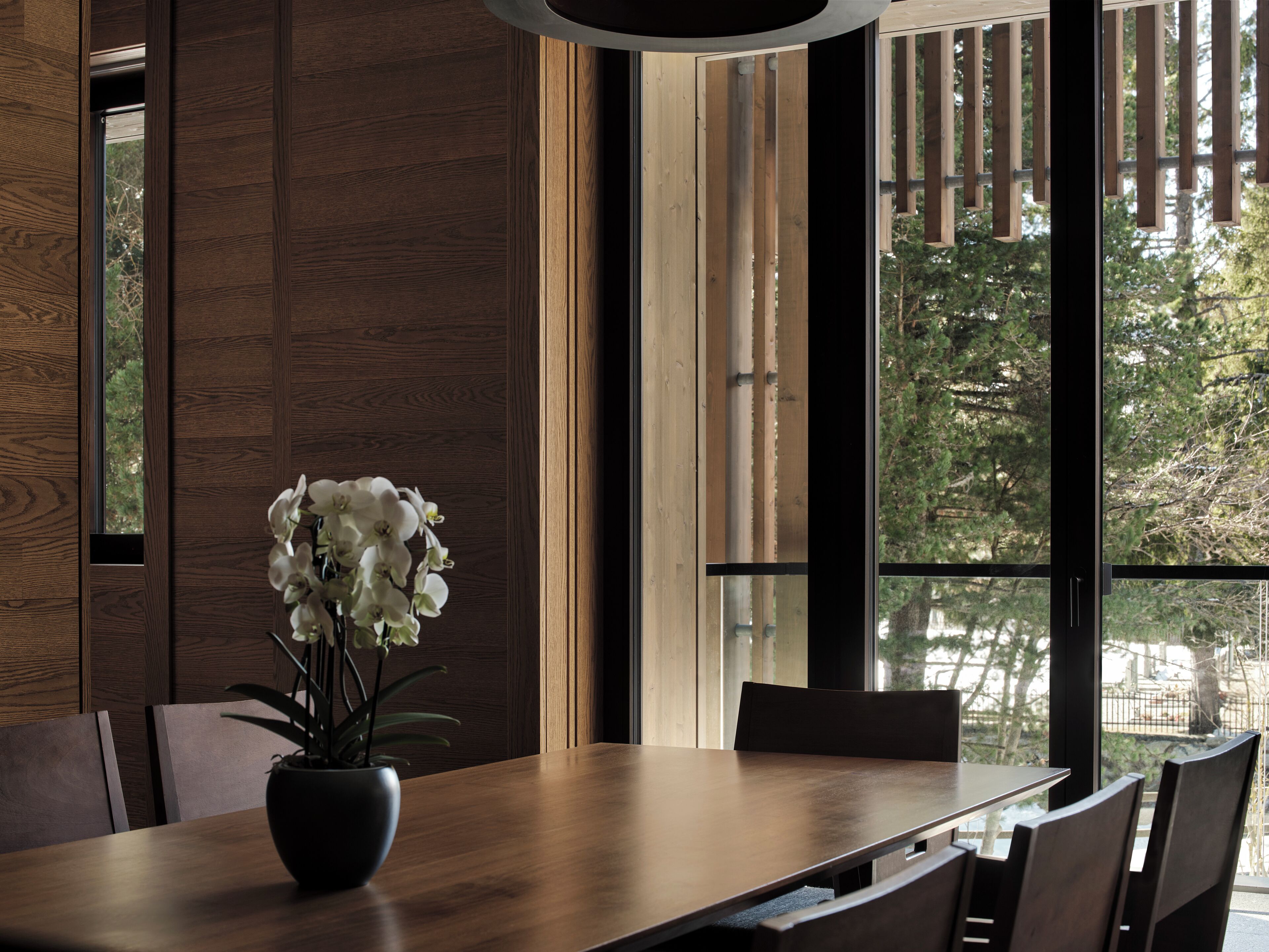 Un espace repas moderne avec une table et des chaises en bois, un vase de fleurs blanches et de grandes fenêtres donnant sur des arbres et une clôture.