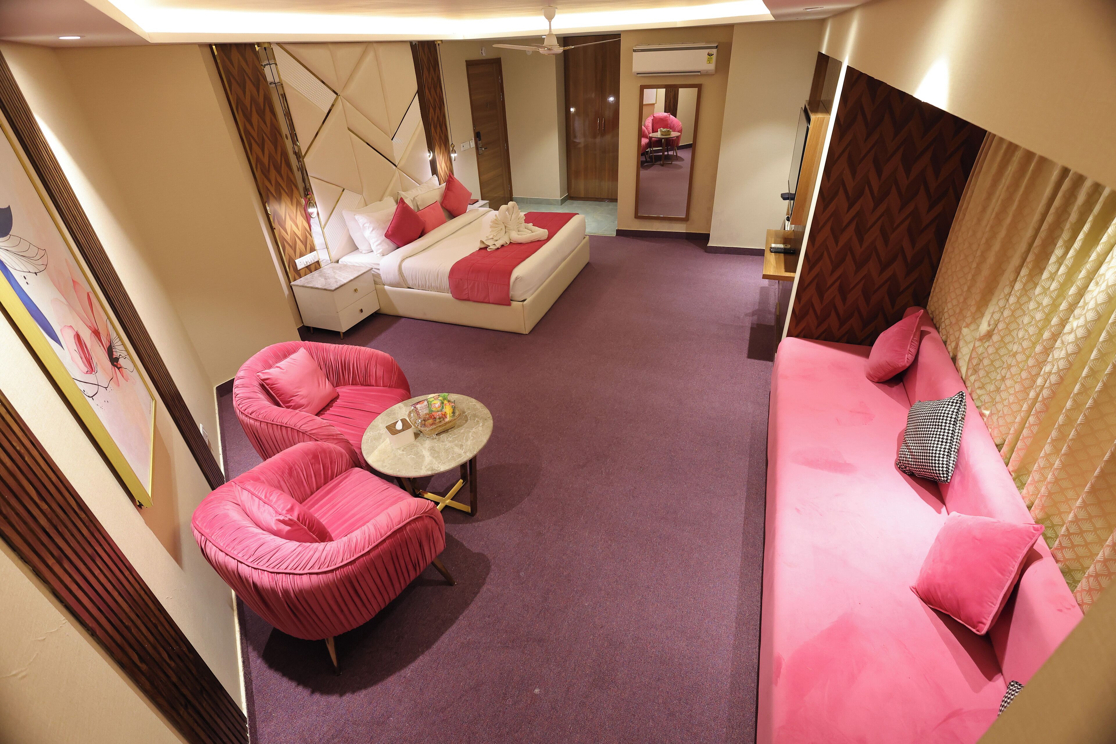 A hotel room with a bed, two pink armchairs, a small round table, and a sofa with a patterned cushion.