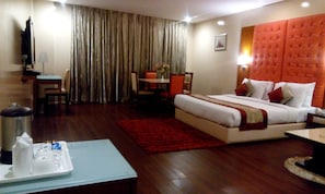 A hotel room with a large bed, a desk with a chair, a dining table with chairs, and a TV. - Hotel Ramhan Palace (New Delhi)